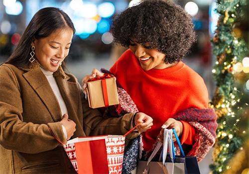 Two female friends are enjoying the Christmas spirit by giving each other presents in a beautifully decorated city street